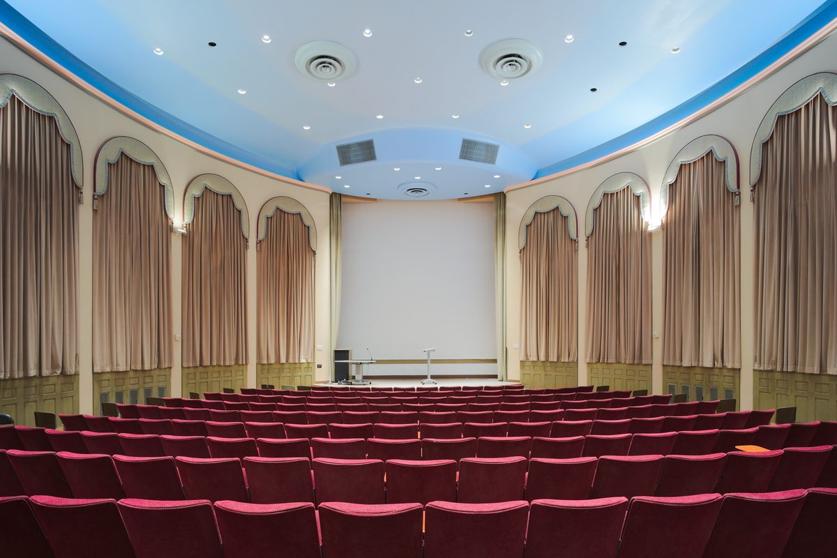 A large panelled university auditorium with tiered seating and a broad stage