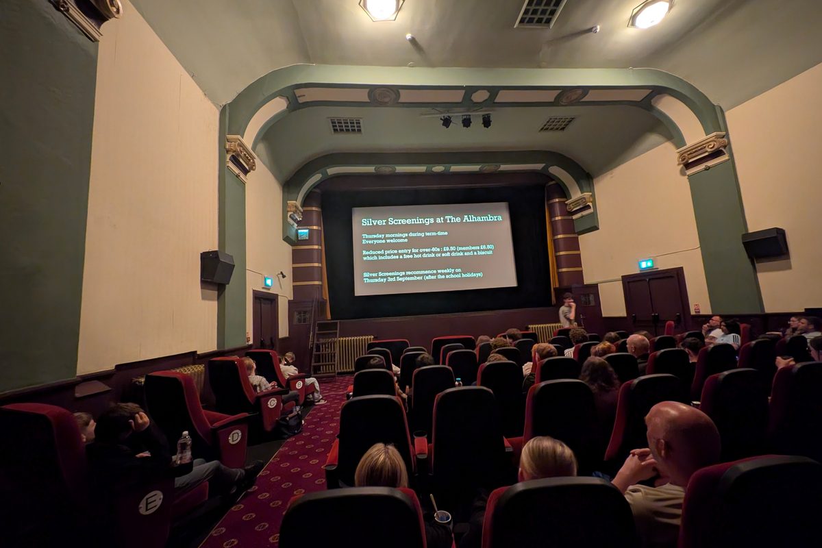 A cinema auditorium with an audience in upholstered rows facing a lit screen under a
      panelled proscenium arch