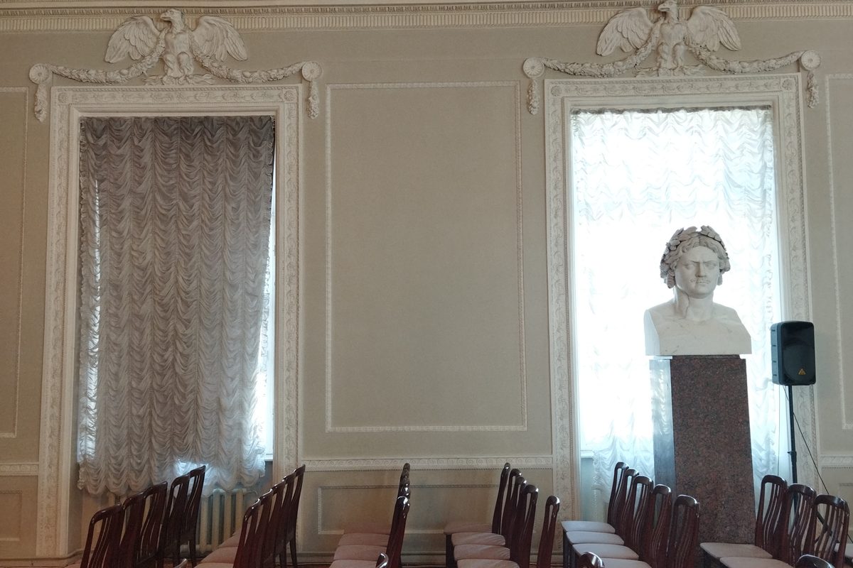 A high-ceilinged institutional conference hall with loose chairs set in rows and tall
      windows along one wall