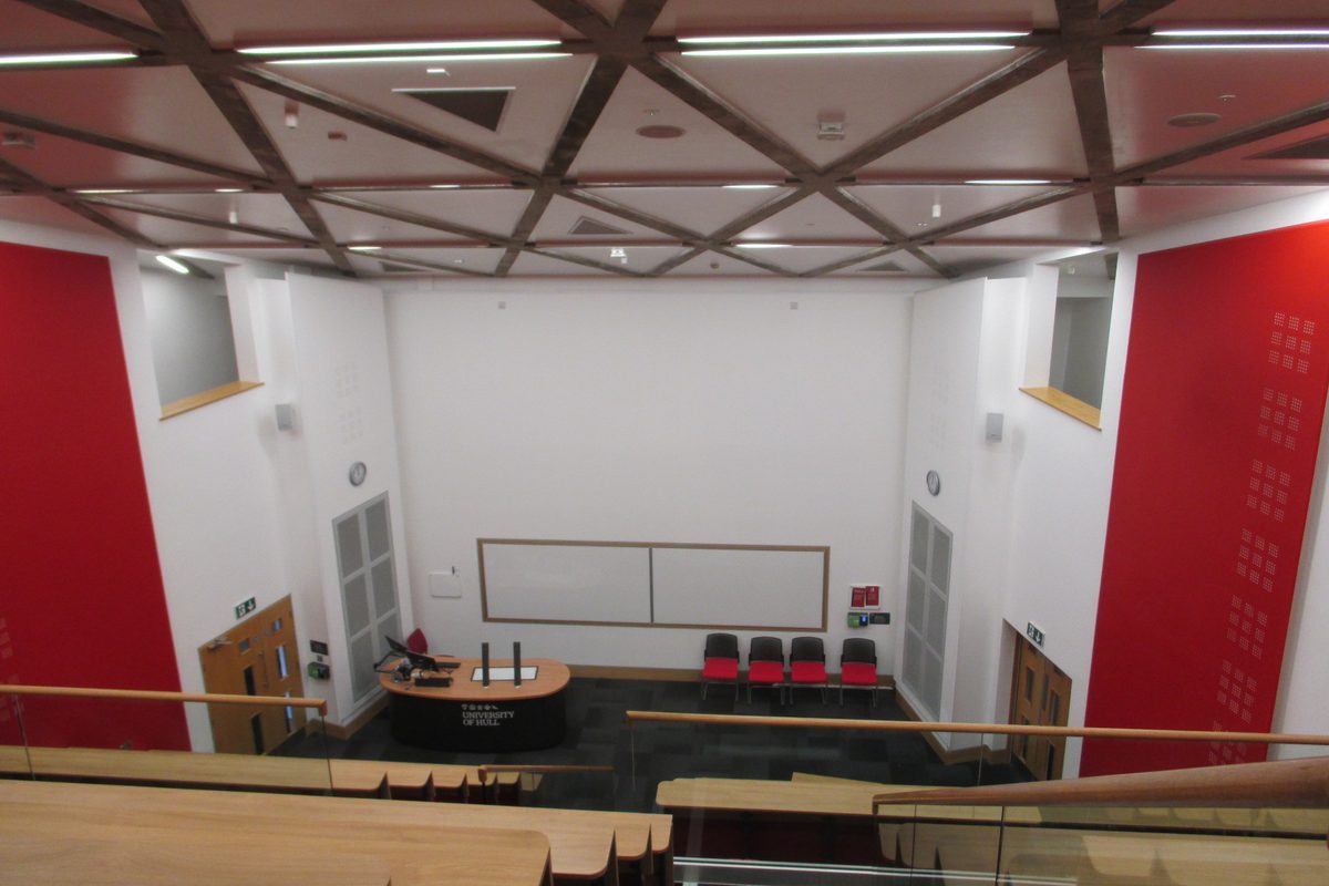 A raked university lecture theatre seen from the back looking down: fixed bench rows
      facing a lectern and a wall-mounted projection surface